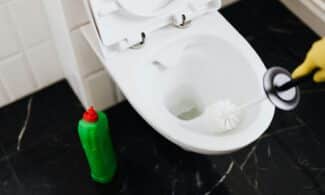 From above of crop anonymous person in rubber gloves using liquid toilet sanitizer and brush to clean toilet bowl in bathroom