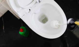 From above crop anonymous person in yellow gloves cleaning toilet bowl with toilet brush and liquid detergent in green plastic bottle