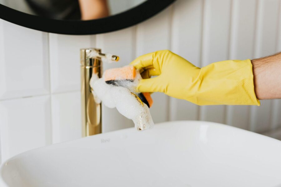 Unrecognizable man wearing yellow latex gloves washing golden faucet in bathroom while cleaning home with orange sponge and detergent