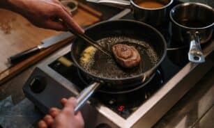 Close-up of pan-seared steak in kitchen stove setting with hands involved, capturing culinary artistry.