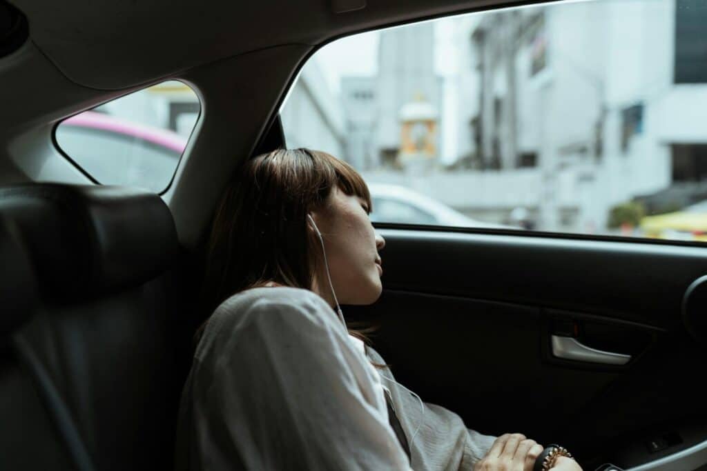 Side view of unemotional female in white shirt resting in car passenger seat and listening to music via earphones while riding in urban environment