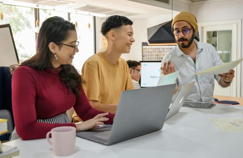 A group of adults engaged in a collaborative work discussion around laptops in a modern office setting.