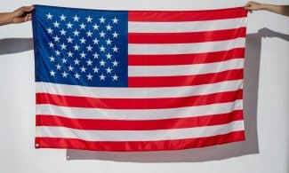 Two hands hold a vibrant American flag against a plain white wall.