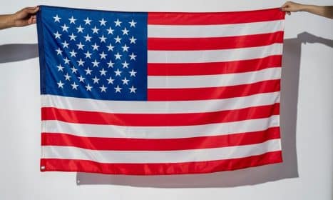 Two hands hold a vibrant American flag against a plain white wall.