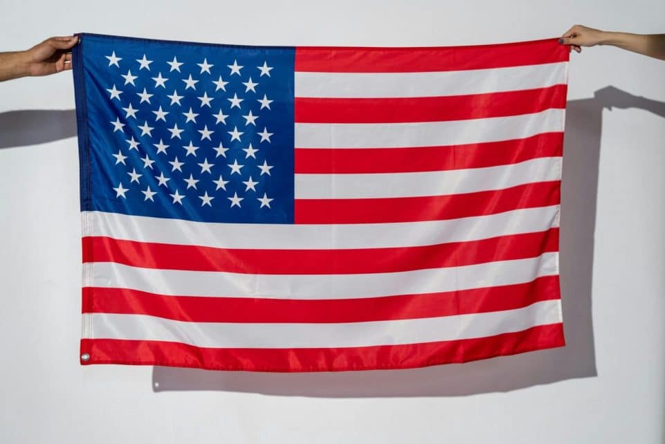 Two hands hold a vibrant American flag against a plain white wall.
