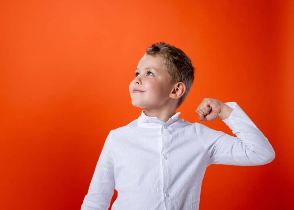 Young boy confidently flexing his arm muscles in a white shirt against a vivid orange background.