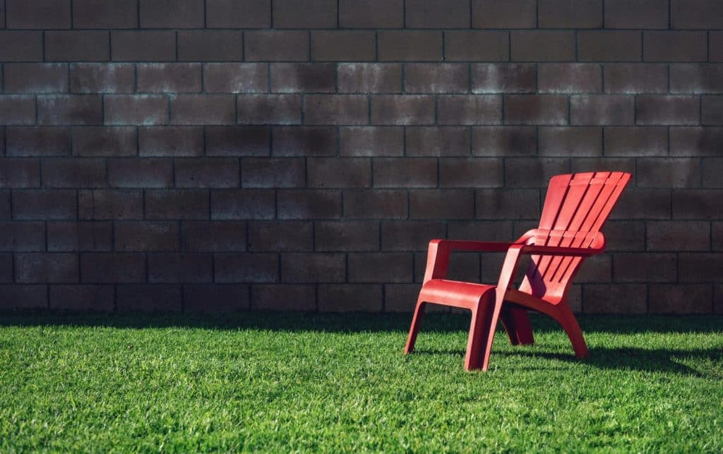 A bright red chair sits on a lush green lawn against a shadowed concrete wall during summer.