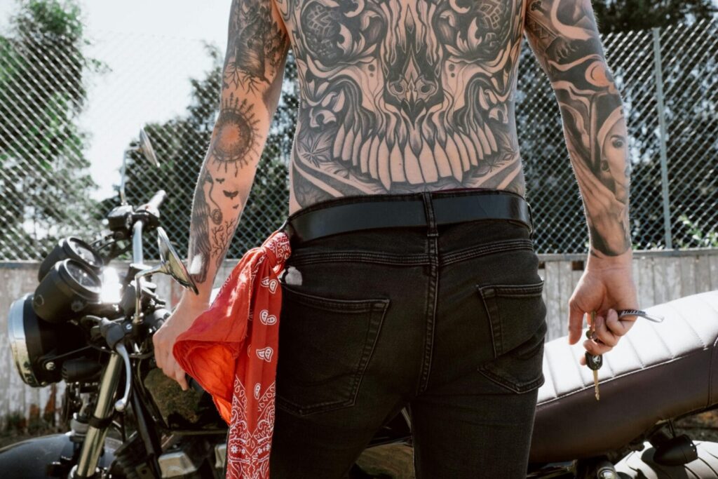 Back view of a tattooed biker holding keys beside a motorcycle, showcasing detailed body art and black jeans.