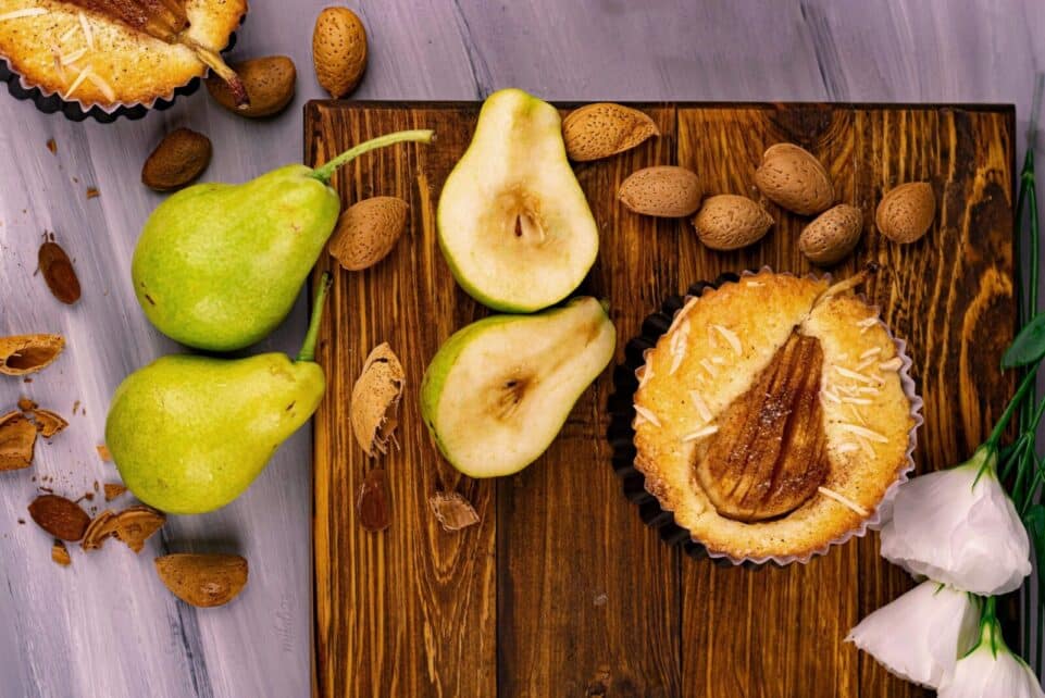 Close-up of a rustic pear tart surrounded by fresh pears and almonds on a wooden board.