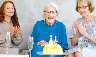 Positive elderly women in stylish clothes sitting on sofa near table with birthday cake and glasses of champagne celebrating birthday at home
