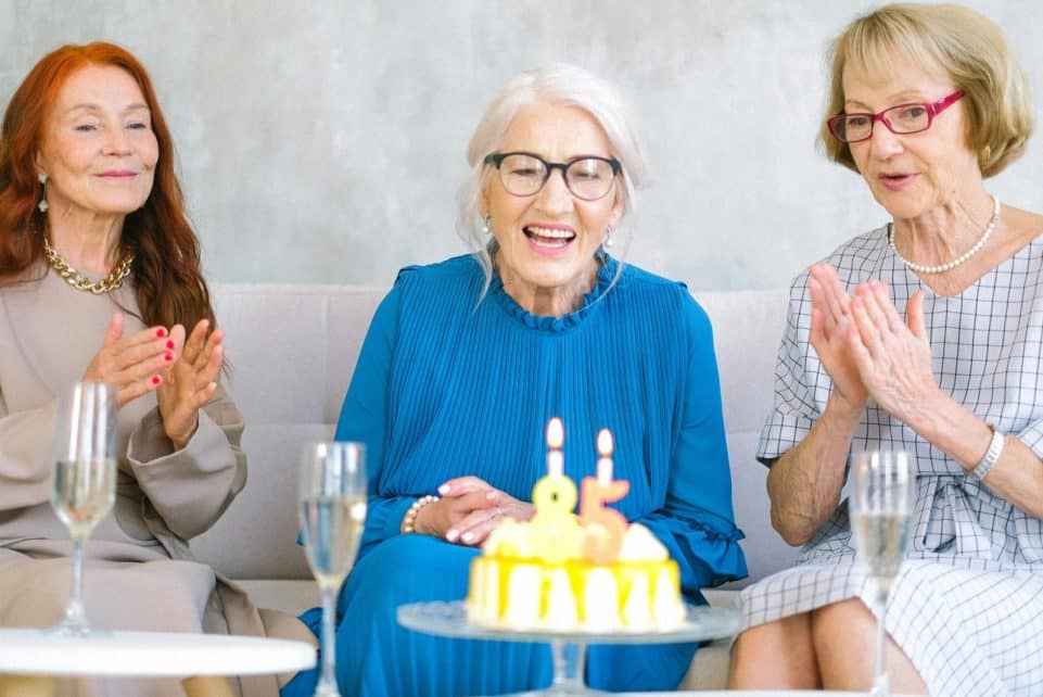 Positive elderly women in stylish clothes sitting on sofa near table with birthday cake and glasses of champagne celebrating birthday at home