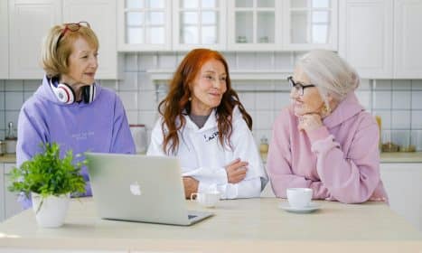 Positive senior women in trendy outfits sitting at table with cups of tea and laptop and communicating in light kitchen at home