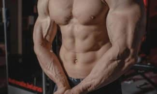 Shirtless male bodybuilder showing muscular torso and defined arms in a gym setting.