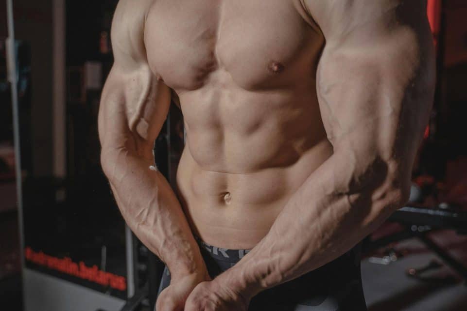 Shirtless male bodybuilder showing muscular torso and defined arms in a gym setting.