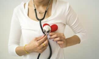 Unrecognizable female wearing white shirt while standing on white background with diaphragm of stethoscope on red handmade heart in room