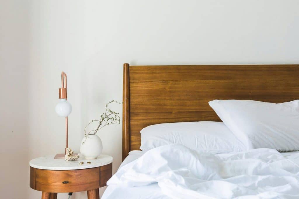A minimalist bedroom features a wooden headboard, white bedding, and a stylish side table lamp.