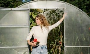 A woman standing at a greenhouse entrance holding produce, embodying autumn harvest.