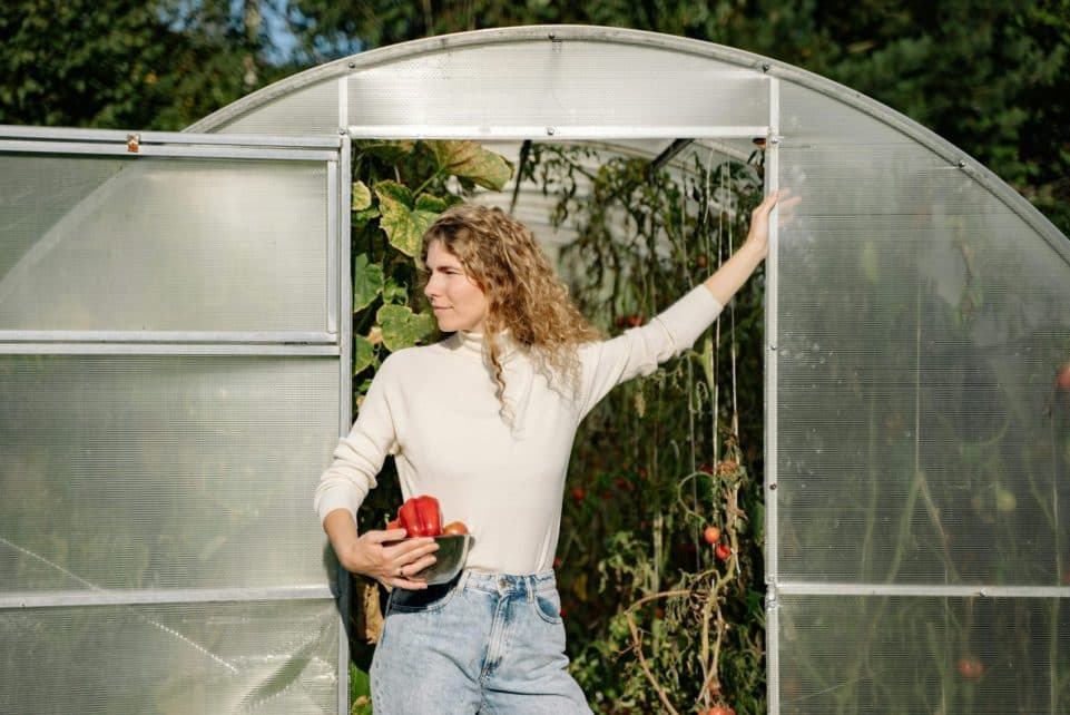 A woman standing at a greenhouse entrance holding produce, embodying autumn harvest.