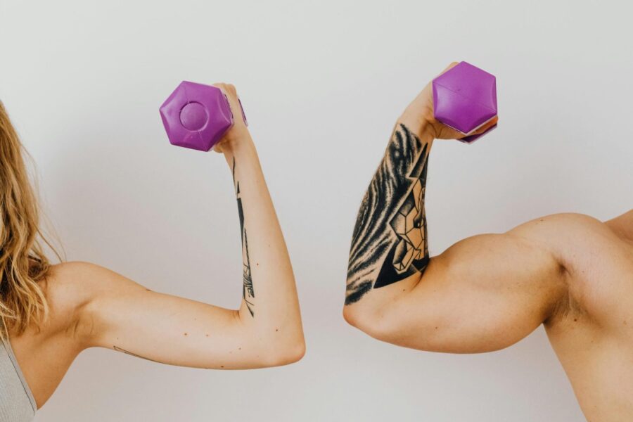 Two individuals flexing their muscles with purple dumbbells, showcasing tattoos and fitness dedication.