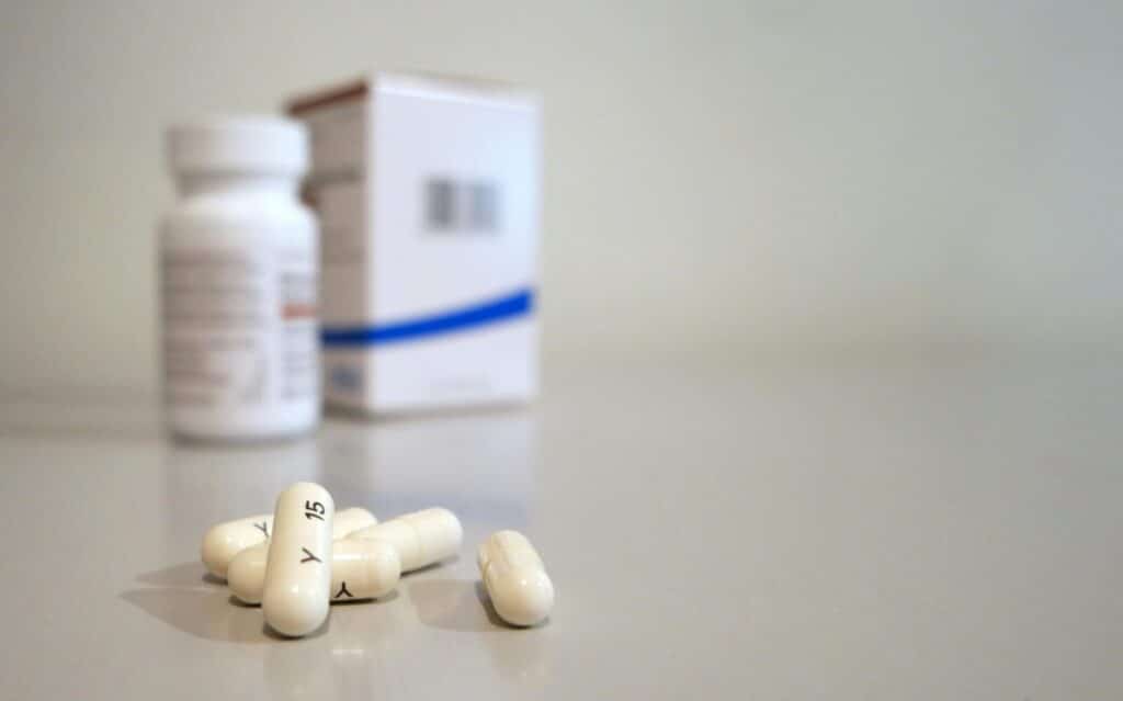 Close-up image of white capsules with medicine bottle and box in the background.