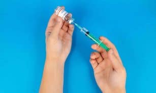 Hands holding a syringe and COVID-19 vaccine vial on a blue background.