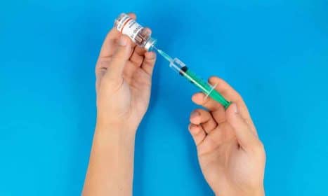Hands holding a syringe and COVID-19 vaccine vial on a blue background.