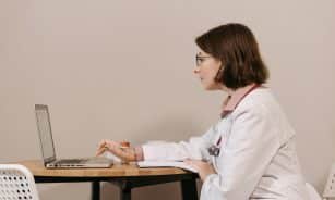 Side profile of a female doctor in a white coat working on a laptop at a desk.