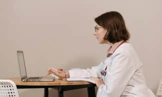 Side profile of a female doctor in a white coat working on a laptop at a desk.