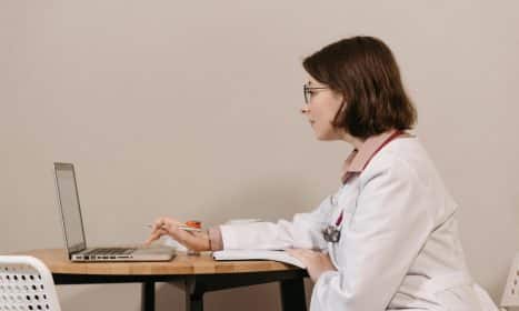 Side profile of a female doctor in a white coat working on a laptop at a desk.