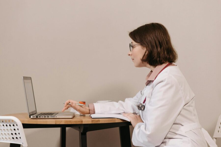 Side profile of a female doctor in a white coat working on a laptop at a desk.