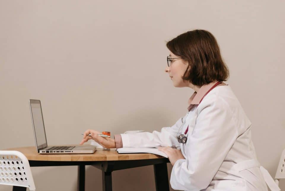 Side profile of a female doctor in a white coat working on a laptop at a desk.