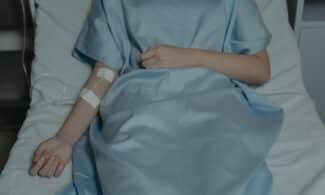 A patient lies in bed wearing a hospital gown with an IV drip attached, evoking a sense of illness.