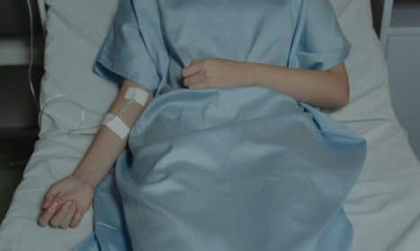 A patient lies in bed wearing a hospital gown with an IV drip attached, evoking a sense of illness.