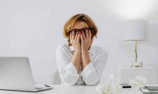 Woman with hands on face, sitting at desk with laptop and tissues, feeling stressed.