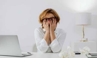 Woman with hands on face, sitting at desk with laptop and tissues, feeling stressed.