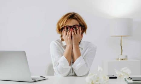 Woman with hands on face, sitting at desk with laptop and tissues, feeling stressed.