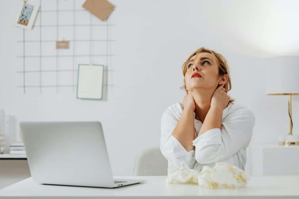 A stressed woman in white long sleeves sits at a desk with a laptop, looking upwards in an office.