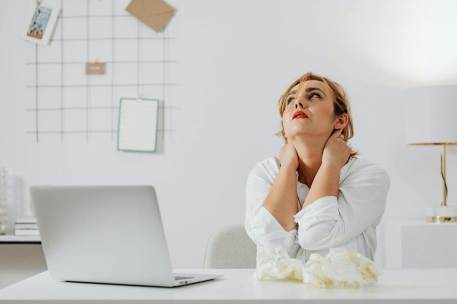 A stressed woman in white long sleeves sits at a desk with a laptop, looking upwards in an office.