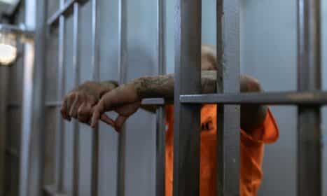 A tattooed inmate's arms rest on jail cell bars, wearing an orange uniform inside a prison.