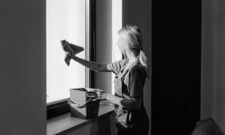 Black and white image of a woman cleaning a window with a cloth and bucket indoors.