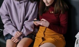 Two teenage girls seated indoors, engrossed in using a smartphone together.