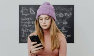 Teenager wearing a beanie and eyeglasses using a smartphone against a blackboard.