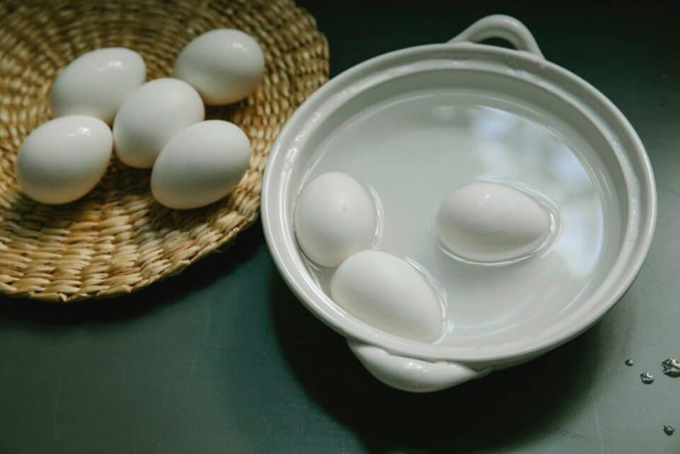 Boiled and raw white eggs arranged in a ceramic bowl and wicker tray on a kitchen surface.