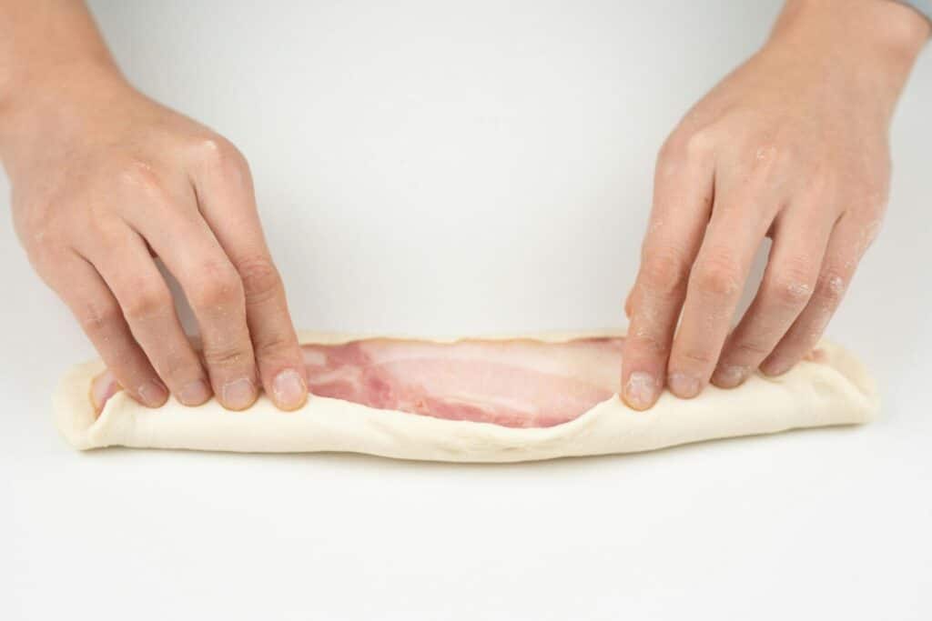 Close-up of hands wrapping bacon in dough on a white background.