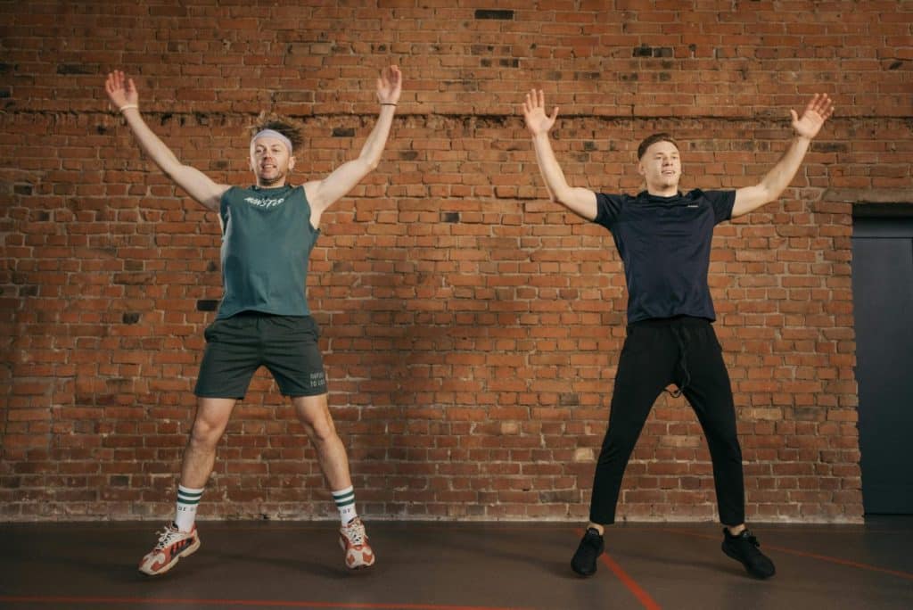 Two men performing jumping jacks in a gym setting, showcasing fitness and energy.
