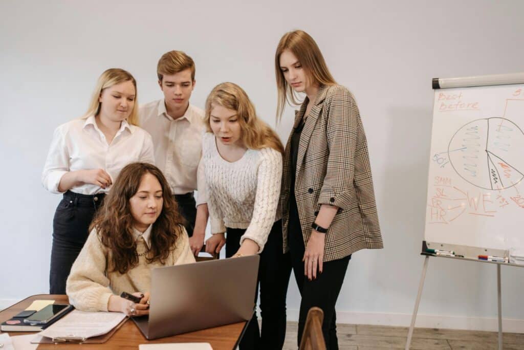 Group of young adults collaborating on a project with a laptop and whiteboard in a modern office setting.