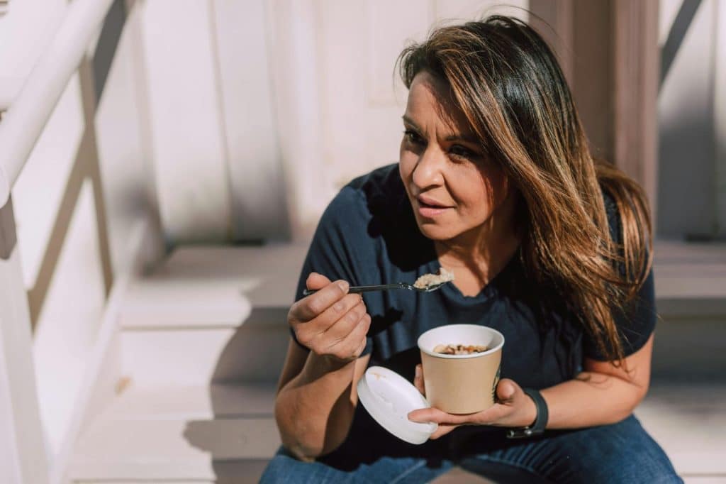 A woman in casual wear eating oats from a disposable bowl while sitting outdoors. Healthy lifestyle concept.