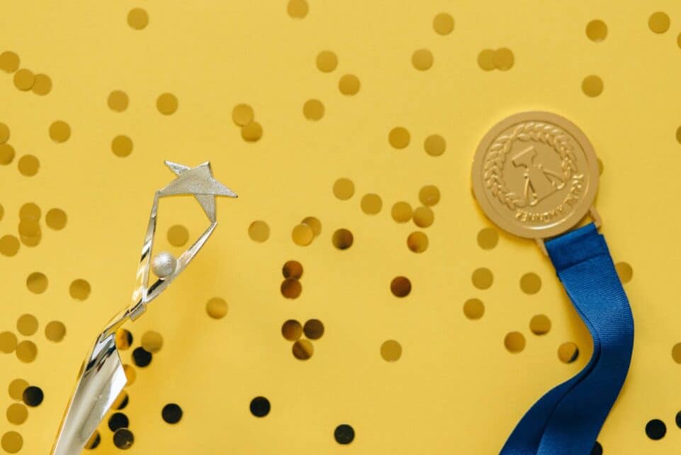 Golden medal and silver trophy with confetti on yellow surface, celebrating achievement.