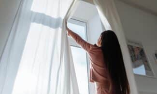 Woman opens white curtains in bright room, revealing a sunny day outside.