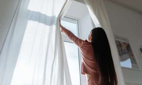 Woman opens white curtains in bright room, revealing a sunny day outside.
